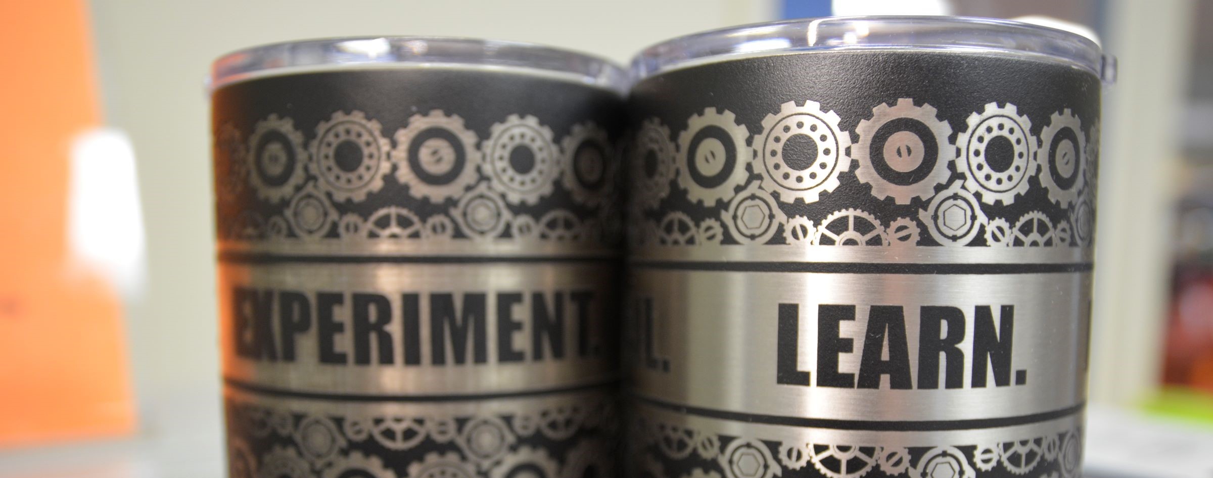 Two lidded tumblers with custom etched pattern of gears and the words "experiment" and "learn" displayed in the Makerspace