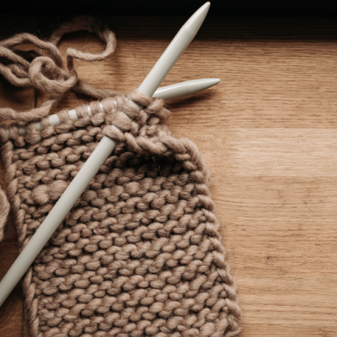 large white knitting needles with the beginning of a tan scarf, sitting on a talble