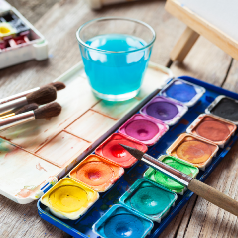 An open watercolor palette with a variety of colors, a cup of water, and paintbrushes.