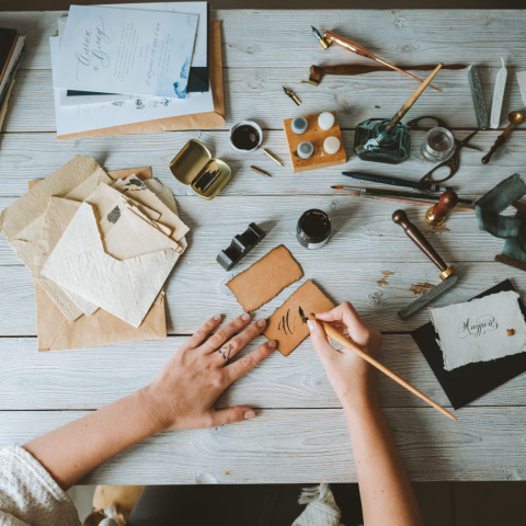 calligraphy supplies laid out on a table with hands holding a pen and writing and a small piece of paper