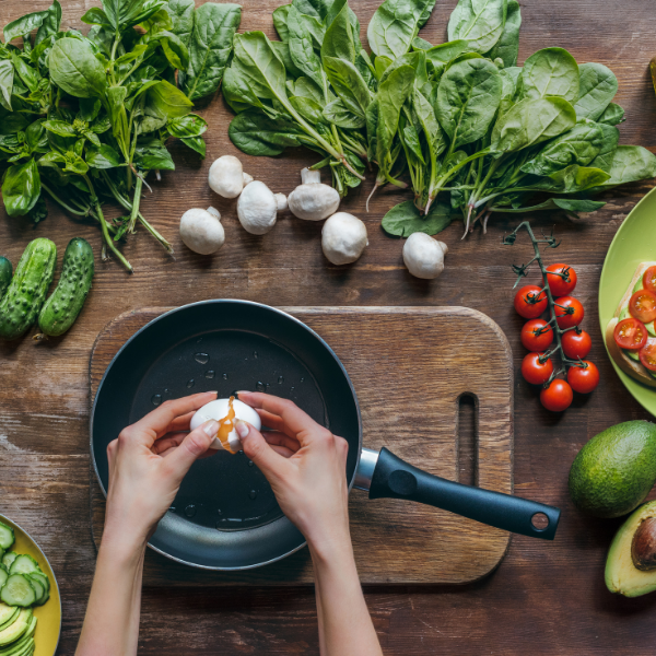 Hands cracking an egg into a pan on a counter covered with vegetables